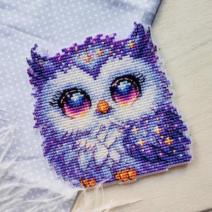 Peut inclure: Une chouette brodée au point de croix, aux yeux colorés et aux ailes étoilées. La chouette présente des nuances de violet, de bleu et de blanc, avec des touches d'orange. Le travail artisanal est détaillé.