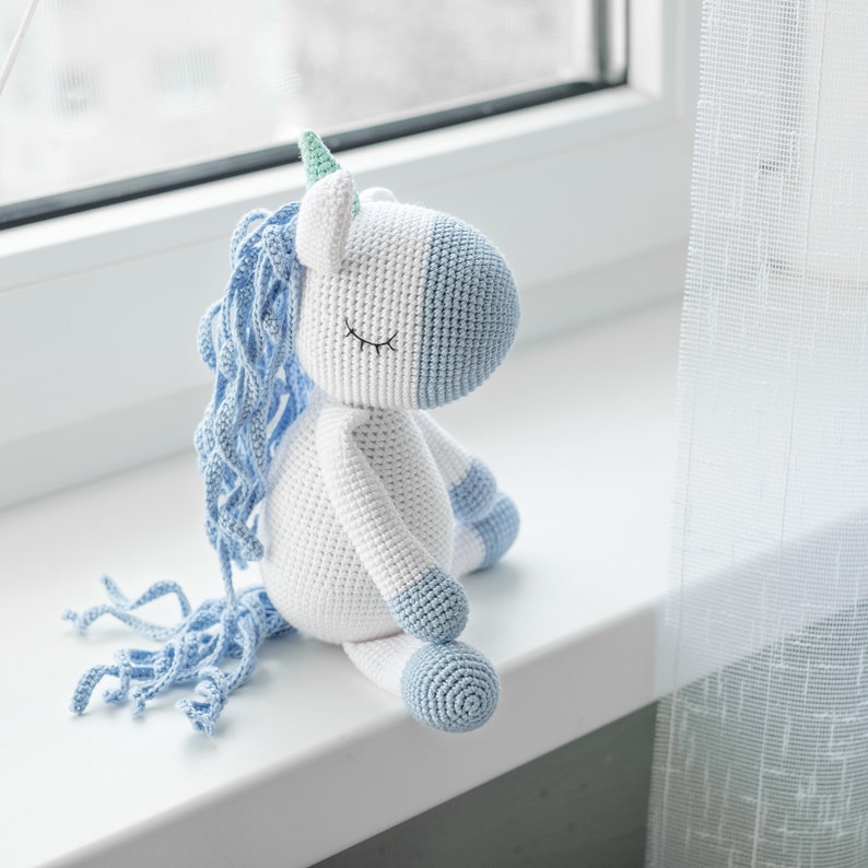May include: A crocheted unicorn toy with a white body, blue mane and tail, and a green horn. The unicorn is sitting on a white surface.