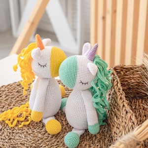 May include: Two crocheted unicorn toys, one with yellow and orange mane and tail, the other with green and purple mane and tail. Both have white bodies and closed eyes.