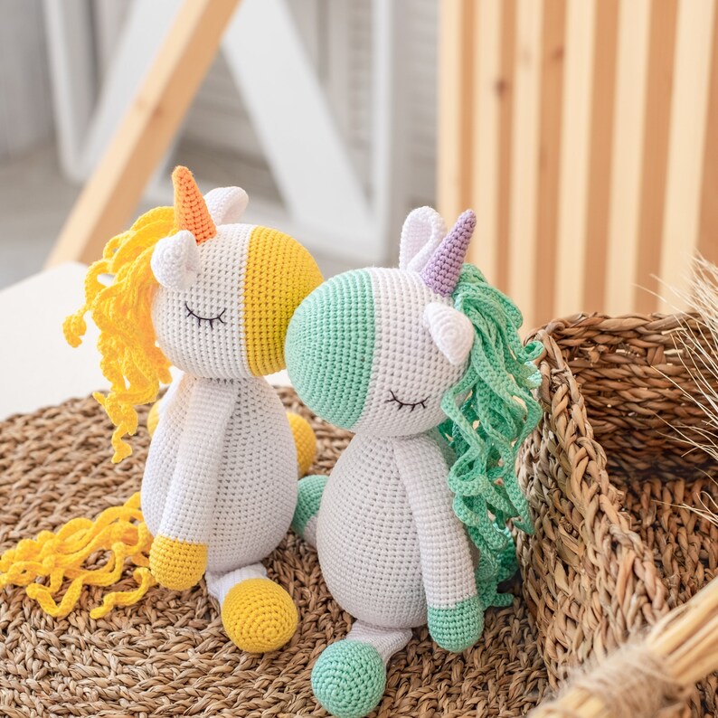 May include: Two crocheted unicorn toys, one with yellow and orange mane and tail, the other with green and purple mane and tail. Both have white bodies and closed eyes.