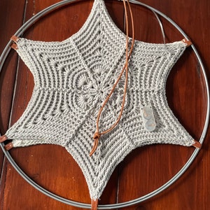 Op de afbeelding: Een grijze gehaakte ster met een bruin leren koord dat eraan hangt. De ster is bevestigd aan een zilveren metalen hoepel.