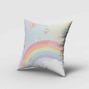 Puede incluir: Una almohada blanca con un arco&iacute;ris pastel y globos aerost&aacute;ticos impresos en ella. El arco&iacute;ris est&aacute; formado por rosa, naranja, amarillo, verde, azul y morado. Los globos aerost&aacute;ticos son amarillos, rosas y azules.