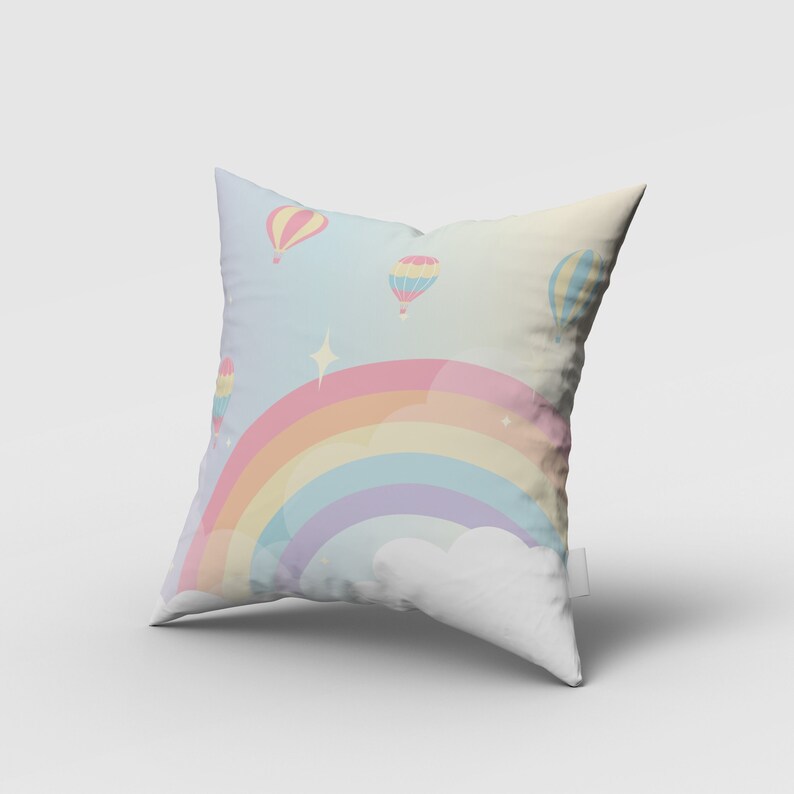 Puede incluir: Una almohada blanca con un arco&iacute;ris pastel y globos aerost&aacute;ticos impresos en ella. El arco&iacute;ris est&aacute; formado por rosa, naranja, amarillo, verde, azul y morado. Los globos aerost&aacute;ticos son amarillos, rosas y azules.
