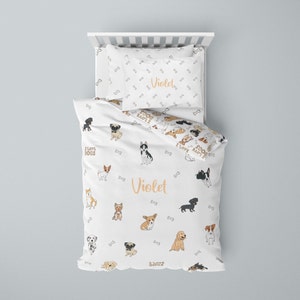Peut inclure: Ensemble de literie blanc avec un motif de chien. La housse de couette et les taies d'oreiller présentent une variété de races de chiens, notamment un carlin, un husky, un teckel et un chihuahua. La literie est personnalisée avec le nom "Violet" en couleur orange clair.