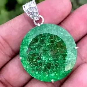 Könnte beinhalten: Ein großer, runder, grüner Smaragd-Anhänger mit einer silbernen Öse. Der Smaragd hat einen facettierten Schliff und eine strukturierte Oberfläche.