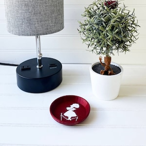 May include: A gray lamp with a black base and a white cord sits on a white surface next to a potted plant with a small figurine inside. A red dish with a white silhouette of a girl is in the foreground.