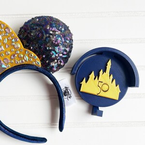 May include: Blue and gold Mickey Mouse ears with a sparkly blue bow and a gold castle charm with the number 50 on it.