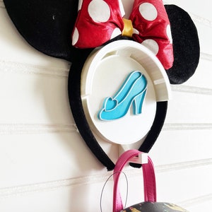 May include: A white headband with black ears and a red bow with white polka dots. The headband has a white plastic charm with a blue glass slipper. A black backpack with a pink zipper and a castle pattern hangs from the headband.