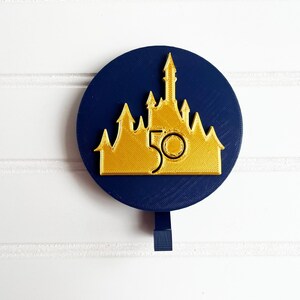 May include: A blue and gold round ornament with a castle silhouette and the number 50. The ornament has a hook on the back.
