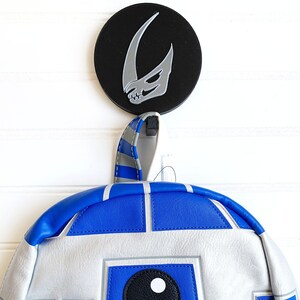 May include: A blue and white R2-D2 backpack with a black and silver Mandalorian symbol on a round black hook.
