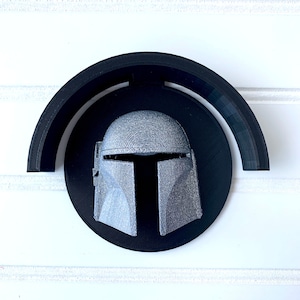 May include: A 3D printed Mandalorian helmet wall mount. The helmet is grey and is mounted on a black circular base with a black arch.