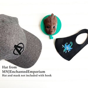 May include: A gray baseball cap with a black Avengers logo, a brown Groot-shaped wall hook, and a black face mask with a blue floral design. The hat and mask are not included with the hook.
