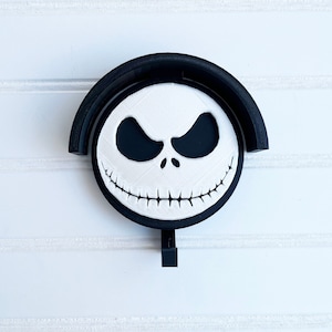 May include: Black and white Jack Skellington face on a round wooden coaster with a black hook. The coaster is attached to a black wooden base.