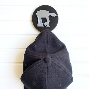 May include: A black and silver AT-AT wall hook with a black baseball cap hanging from it. The cap has a yellow "Star Wars" logo on the front.