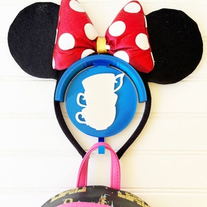 May include: A black headband with red and white polka dot Minnie Mouse ears and a blue and white teacup charm. The headband is hanging on a white wall with a black and pink backpack hanging below it.