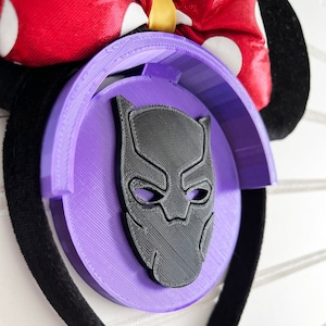 May include: A purple and black ear holder with a Black Panther mask design. The holder is attached to a black headband with a red bow with white polka dots.