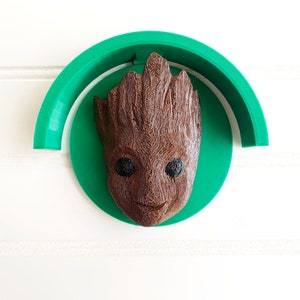 May include: A brown, textured Groot head sculpture with black eyes and a smiling expression, mounted on a green circular base and a green arch. The sculpture is set against a white background.