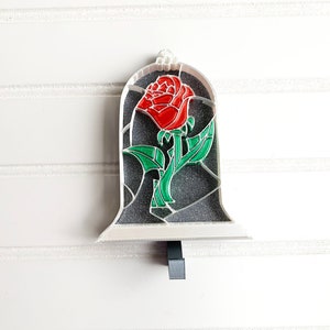 May include: A white ceramic ornament shaped like a glass dome with a red rose inside. The rose has green leaves and a stem. The ornament has a hook on the bottom for hanging.