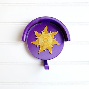 Pode incluir: Um porta-copos roxo montado na parede com um design de sol dourado. O suporte tem uma base circular com uma parte superior curva e um pequeno gancho na parte inferior. O design do sol é centrado na base e tem uma aparência texturizada detalhada.