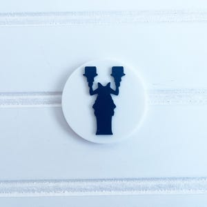 Peut inclure: Un emblème blanc et circulaire représentant une silhouette bleu foncé de la Statue de la Liberté. L'emblème est centré sur une surface blanche texturée avec des lignes horizontales.