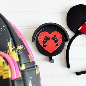 May include: A black backpack with a pink zipper and gold trim features a pattern of gold castles. A black headband with a red and white polka dot bow and black ears. A black and red heart-shaped phone holder with a silhouette of Mickey and Minnie Mouse kissing.