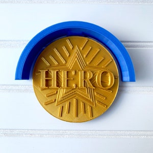 May include: A gold-colored, 3D printed, round plaque with a star and the word "HERO" in raised letters. The plaque is sitting inside a blue plastic ring.