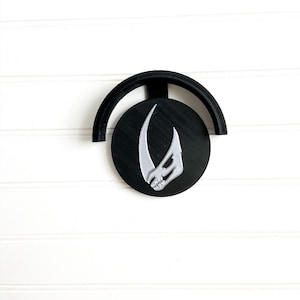 May include: A black and white Mandalorian symbol wall mount. The symbol is a curved, stylized skull with a horn.