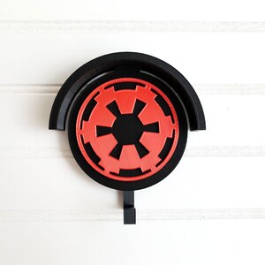 May include: A black wall hook with a red and black Imperial symbol from Star Wars. The hook is shaped like a half circle with a hook extending from the bottom.