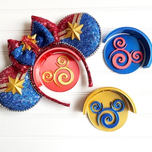 May include: A pair of red, white, and blue sequined Minnie Mouse ears with a gold star on each ear. The ears are on a red headband with a gold Mickey Mouse icon on the headband. There are two additional Mickey Mouse ear stands, one blue and one gold.