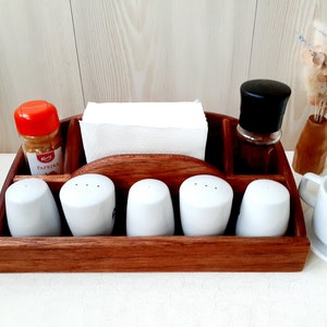 Può includere: Un portacondimenti in legno con un portatovaglioli integrato, cinque saliere in ceramica bianca e due barattoli di spezie. Un barattolo di spezie ha un coperchio arancione e l'altro un coperchio nero. Il portacondimenti è in legno scuro.