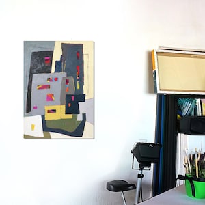 May include: Abstract cityscape painting with geometric shapes in gray, yellow, red, and blue. The artwork hangs on a white wall, with a studio setting visible in the background, including canvases and art supplies.