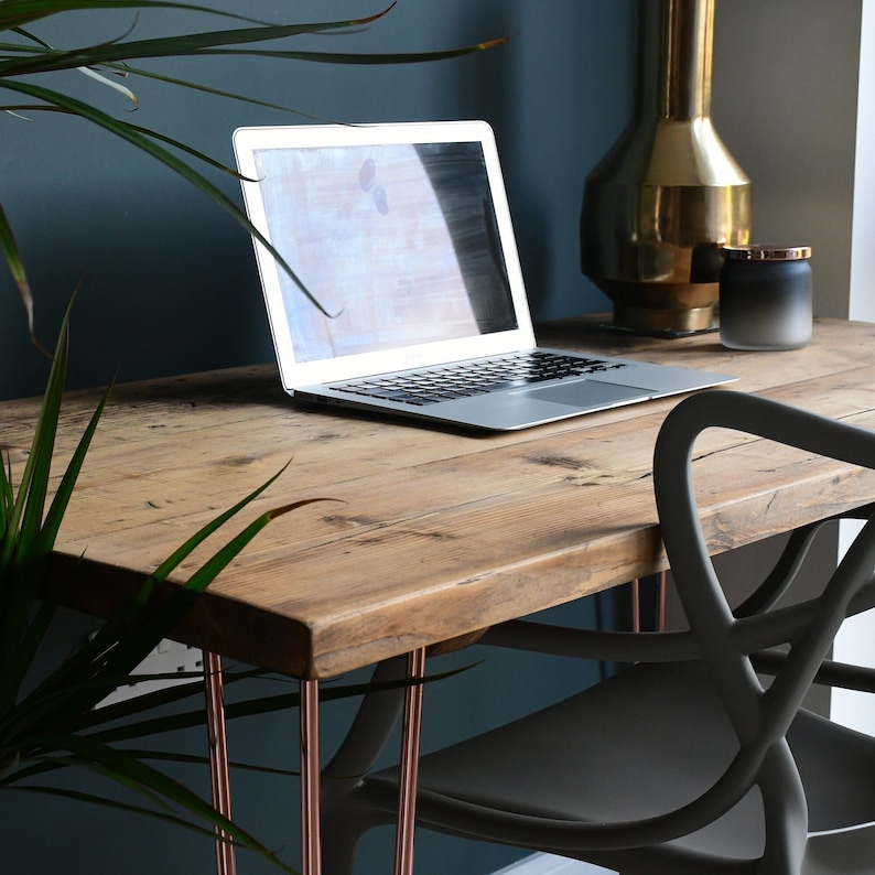 May include: A rustic wooden desk with copper hairpin legs. A laptop computer is on the desk, and a black chair is in the foreground. The desk is against a teal wall.