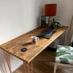 May include: A rustic wooden desk with copper hairpin legs. The desk has a laptop, two mugs, and a lamp on it. The desk is in a room with a white wall and a patterned chair.