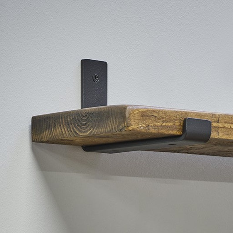 Op de afbeelding: Een rustieke houten plank met een donkere afwerking, ondersteund door twee zwarte metalen beugels. De plank is gemaakt van dik, verweerd hout, met natuurlijke nerfpatronen. De beugels zijn bevestigd aan een lichtgrijze muur, wat contrasteert met het hout. Ongeveer 30 cm diep.