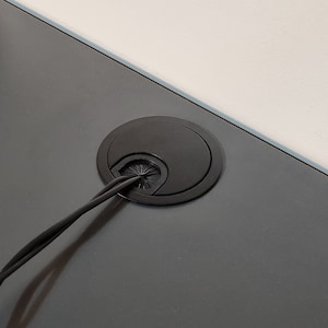 Puede incluir: Grommet de gesti&oacute;n de cables negro instalado en una superficie gris oscuro. El grommet tiene una abertura circular con una cubierta de pl&aacute;stico negro.