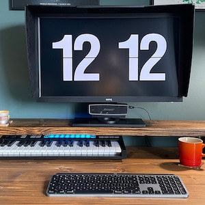 May include: A wooden monitor stand with a keyboard, a keyboard, a mug, and a candle on a wooden desk. The monitor displays the time "12 12".
