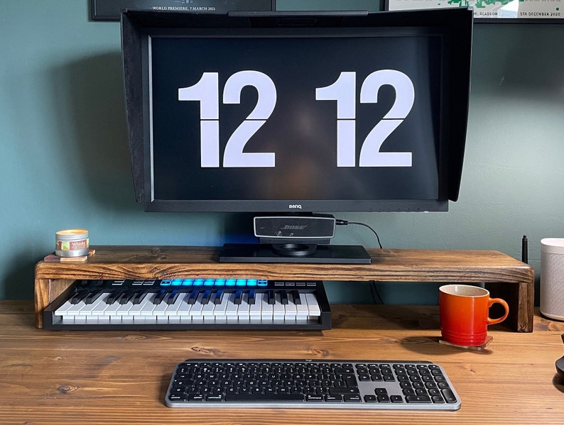 May include: A wooden monitor stand with a keyboard, a keyboard, a mug, and a candle on a wooden desk. The monitor displays the time "12 12".