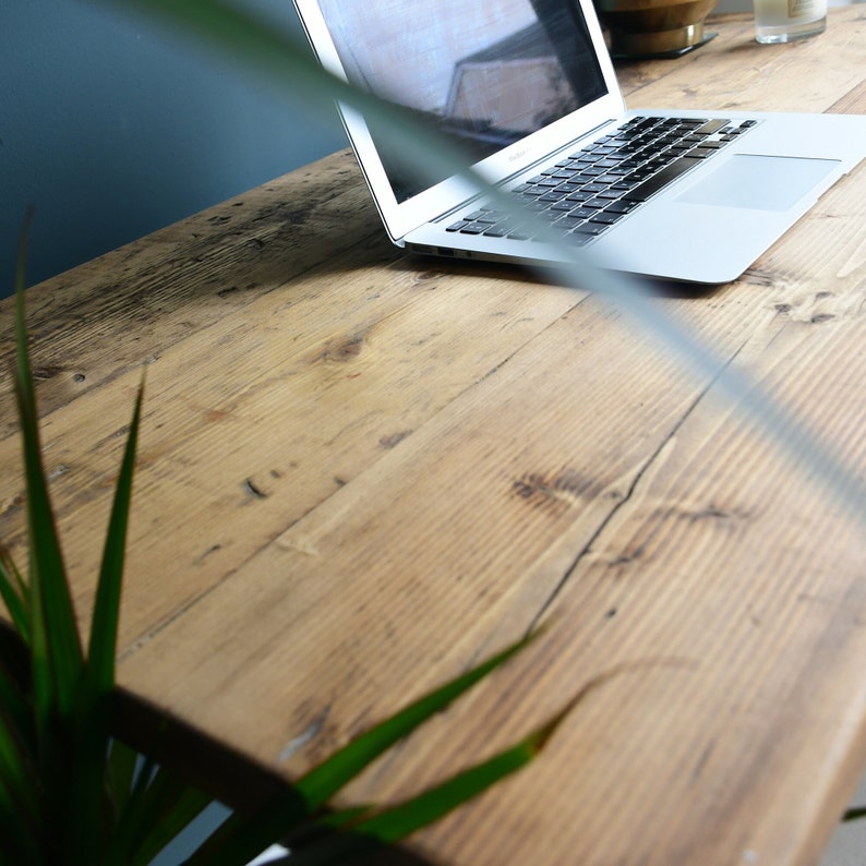 May include: A close-up of a rustic wooden desk with a laptop computer on the surface. The desk is made of reclaimed wood with a natural finish. The laptop is closed and has a silver finish.