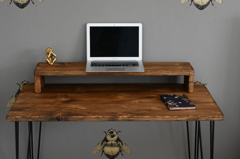 May include: A dark brown wooden desk with black metal hairpin legs. The desk has a built-in laptop stand and a small gold figurine on the top shelf. A notebook with a floral pattern is on the desk.