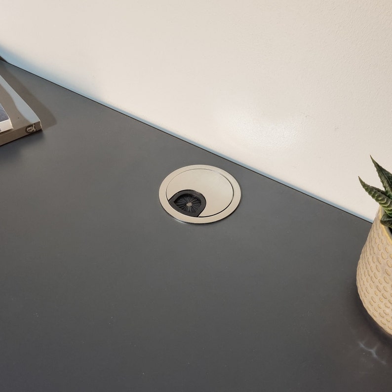 Puede incluir: Primer plano de un pasacables de escritorio plateado con un inserto de goma negro. El pasacables est&aacute; instalado en una superficie gris oscuro.