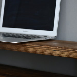 May include: A laptop computer sits on a dark brown wooden shelf. The shelf has a natural wood grain and is made of solid wood.