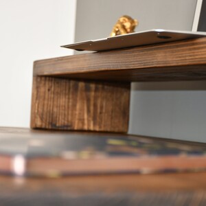 May include: A wooden laptop stand with a dark brown stain. The stand is raised on a single, thick, rectangular piece of wood. A laptop is sitting on the stand with a small gold figurine on top of it. A book is sitting on a wooden surface below the stand.
