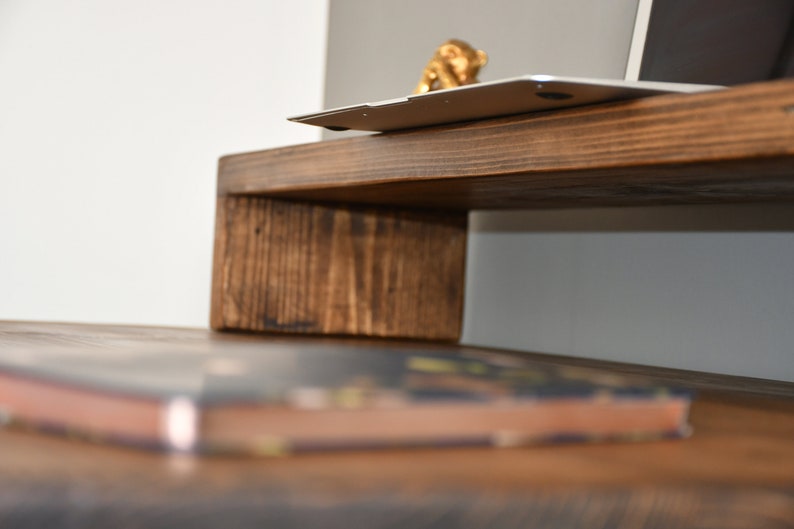 May include: A wooden laptop stand with a dark brown stain. The stand is raised on a single, thick, rectangular piece of wood. A laptop is sitting on the stand with a small gold figurine on top of it. A book is sitting on a wooden surface below the stand.