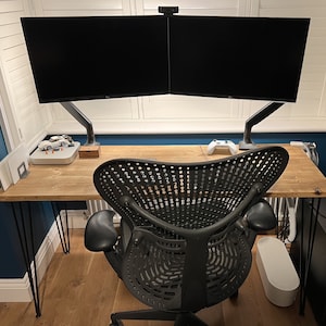 May include: A wooden desk with black metal legs, a black ergonomic office chair, and two black computer monitors mounted on a dual-arm stand. The desk has a white webcam on top of the monitors.