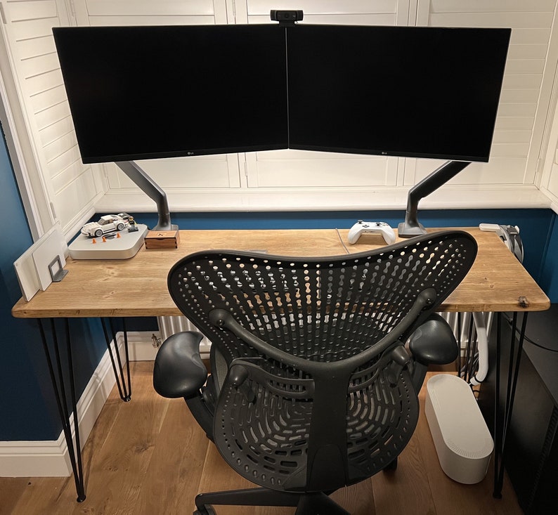 May include: A wooden desk with black metal legs, a black ergonomic office chair, and two black computer monitors mounted on a dual-arm stand. The desk has a white webcam on top of the monitors.