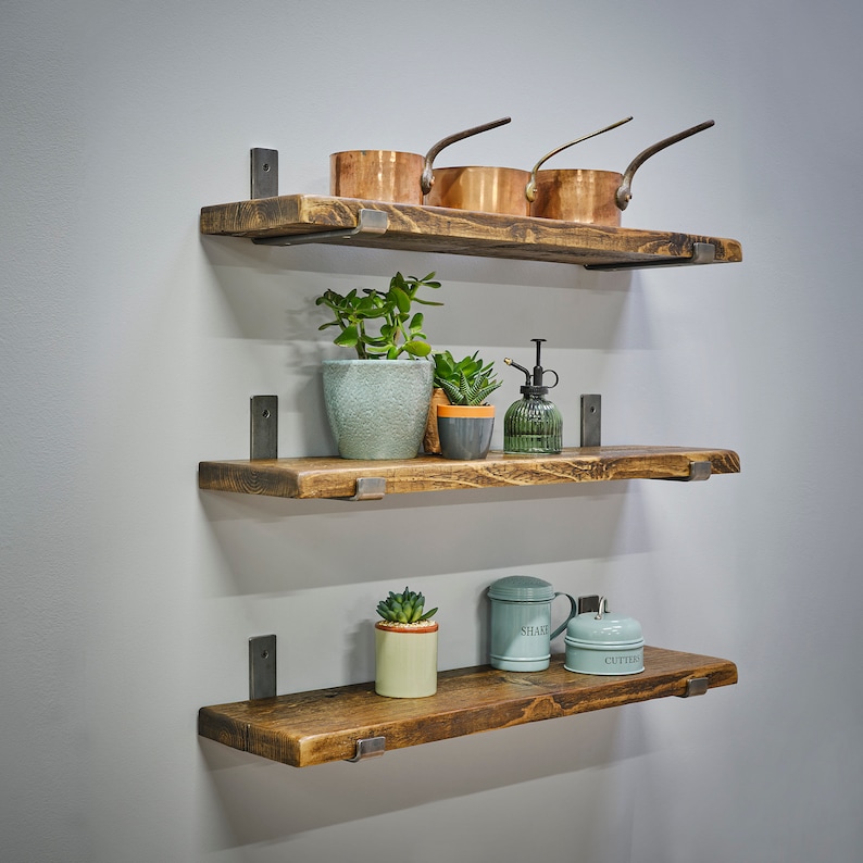 Op de afbeelding: Drie rustieke houten planken met metalen beugels aan een grijze muur. Ze tonen koperen pannen, potplanten en decoratieve containers. Het hout heeft een natuurlijke, verweerde uitstraling, wat een vleugje landelijke stijl toevoegt.