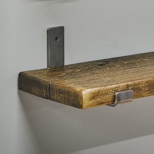 Op de afbeelding: Een rustiek houten plank met een natuurlijke houtnerf en een warme bruine kleur, ondersteund door twee zilveren metalen beugels. De plank is bevestigd aan een lichtgrijze muur en toont een eenvoudig, industrieel ontwerp.
