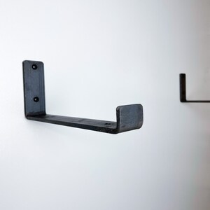 May include: Two black metal shelf brackets mounted on a white wall. The brackets are L-shaped with a flat surface on the top and a curved edge on the bottom.