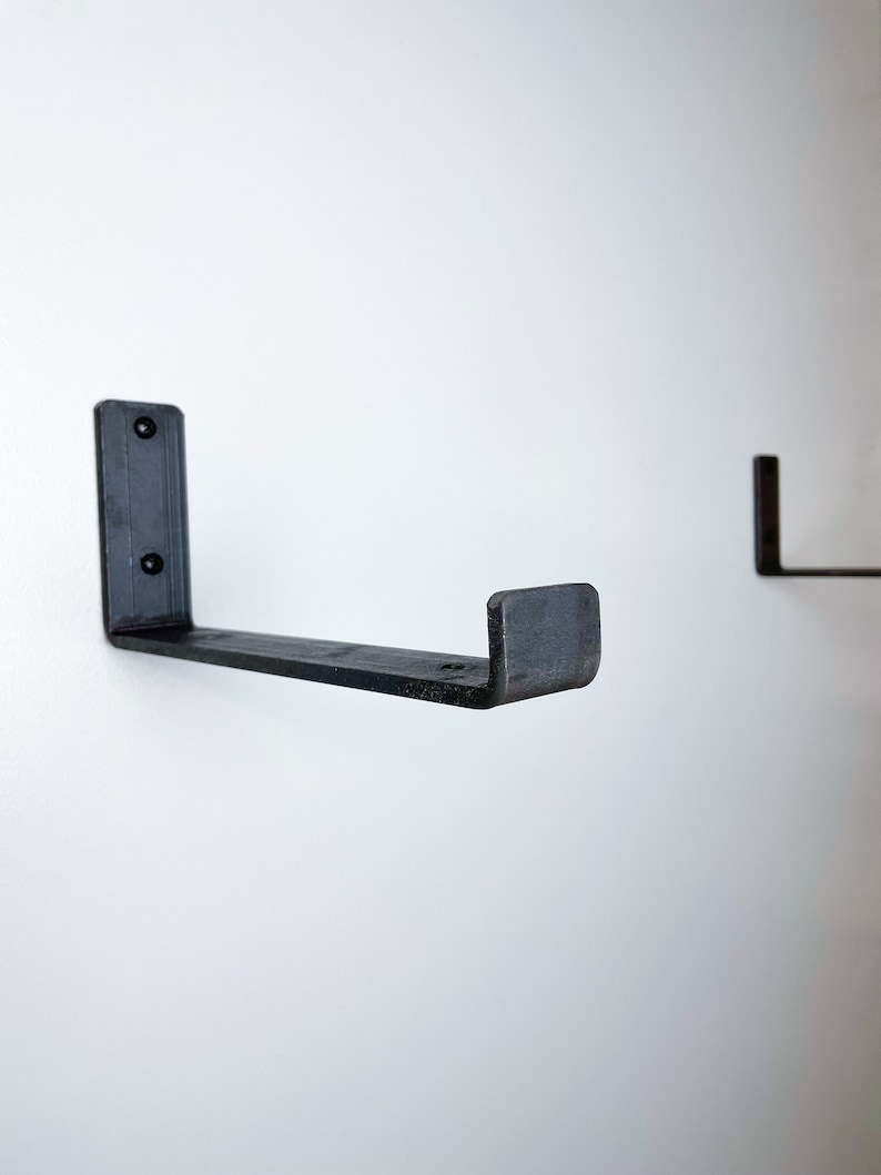 May include: Two black metal shelf brackets mounted on a white wall. The brackets are L-shaped with a flat surface on the top and a curved edge on the bottom.