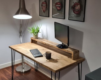 May include: A wooden desk with black hairpin legs, a monitor, and a keyboard. A wooden monitor stand holds a potted plant. A silver lamp and artwork are in the background. The desk has a natural wood finish.